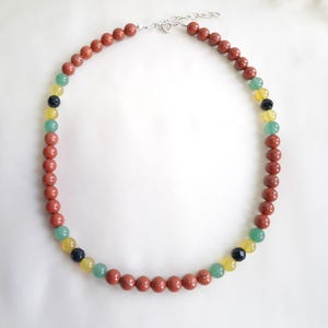 May include: A beaded necklace featuring reddish-brown, green, yellow, and black beads. The necklace has a silver-colored clasp and is displayed on a white surface. The beads are round and evenly spaced.