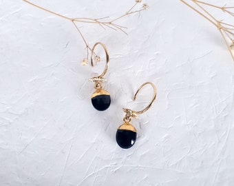 May include: A pair of gold-colored hoop earrings with a black, oval-shaped stone pendant. The earrings have a small, decorative element near the hoop and are displayed on a white textured surface.