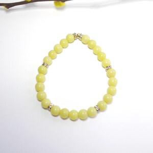 Puede incluir: Una pulsera de cuentas amarillo claro con detalles dorados y espaciadores de diamantes de imitación. La pulsera está hecha de cuentas redondas y lisas. Una pequeña rama con capullos amarillos está en el fondo.