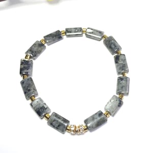 May include: A bracelet made of rectangular grey and black stones, linked by gold-coloured metal beads. The bracelet is presented on a white surface, highlighting its design and colour contrast. The bracelet is a piece of jewellery.