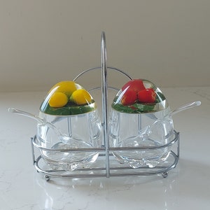 Può includere: Un set di due barattoli per condimenti in vetro trasparente con coperchi che presentano frutta finta. I barattoli sono tenuti insieme da un supporto in filo cromato. I coperchi presentano un limone giallo e un pomodoro rosso.