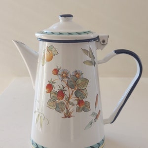 Pode incluir: Bule de esmalte branco com borda azul e um desenho floral com morangos, folhas e uma pêra. A chaleira tem uma tampa com uma pega azul e uma alça.