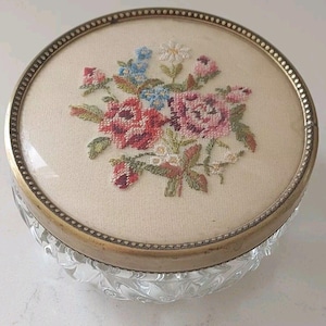 Könnte beinhalten: Ein runder, klarer Glasbehälter mit einem dekorativen, geschliffenen Muster auf der Basis. Der Deckel hat einen cremefarbenen Stoff, der mit einem Blumendesign in Rot-, Rosa-, Blau- und Grüntönen bestickt ist. Der Deckel ist mit einem goldfarbenen Metallband eingefasst.
