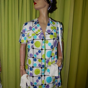 May include: A vintage dress featuring a white base with a geometric pattern of blue, green, and purple circles and squares. The dress has a green zip, short sleeves, and a matching headband. The mannequin also wears sunglasses and white gloves.