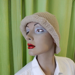 May include: A beige knitted hat with a brim, shown on a mannequin. The hat has a textured appearance and a slightly darker trim. The mannequin is wearing a white shirt, with green curtains in the background.
