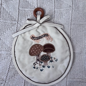 May include: A round, off-white fabric potholder featuring an embroidered design of three brown mushrooms and the text "MUSHROOMS" in a banner. The item includes a dark brown fabric trim, a fabric tie at the top, and a small wooden ring for hanging.
