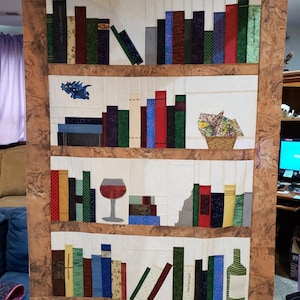 May include: A fabric bookshelf quilt with a brown marbled border. The quilt features shelves with colorful book designs, a wine glass, a basket of flowers, and a bottle. The quilt is a unique, handmade piece.