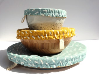 Może przedstawiać: Trzy miski z pokrywkami z tkaniny. Górna miska jest biała z pokrywką w niebiesko-biały wzór. Środkowa miska jest wykonana z naturalnego drewna z pokrywką w żółto-biały wzór. Dolna miska jest wykonana z naturalnego drewna z pokrywką w niebiesko-biały wzór.