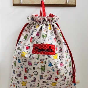 Könnte beinhalten: Ein Kordelzugbeutel mit weißem Hintergrund, der mit farbenfrohen Cartoon-Monster-Illustrationen bedeckt ist. Die Tasche hat einen roten Kordelzug und einen roten Tupfenfleck mit dem aufgestickten Namen "Mamale". Die Tasche hängt an einem hölzernen Wandhaken.
