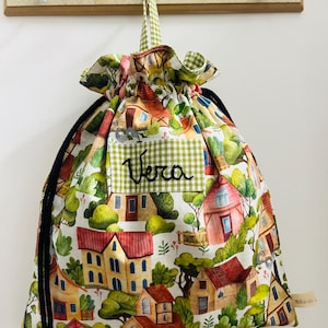 May include: A drawstring bag featuring a colorful village scene with red-roofed houses and lush greenery. The bag has a green gingham trim and the name "Vera" embroidered on the front. Black drawstring closure.