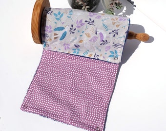 Op de afbeelding: Een paars-wit gepatroneerd keukendoek met een bloemenmotief en vogels. De handdoek is opgerold op een houten stok en heeft een blauwe rand.