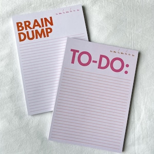 Peut inclure: Deux blocs-notes blancs avec des pages lignées. L'un porte les mots "BRAIN DUMP" en orange, et l'autre "TO-DO:" en rose. Les deux blocs-notes ont les jours de la semaine imprimés en haut en petits caractères.