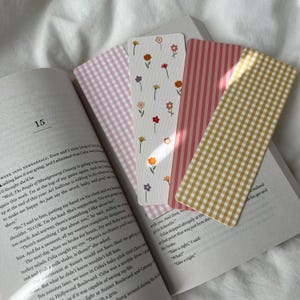 May include: Four decorative bookmarks rest on an open book. The bookmarks feature various patterns: pink gingham, floral, pink stripes, and yellow gingham. The book's page number 15 is visible.