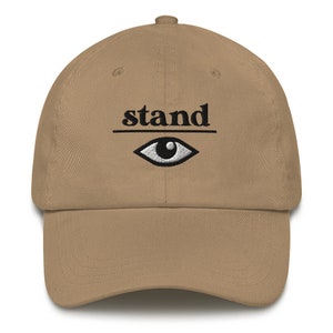 May include: A khaki baseball cap with black embroidery that reads "stand" with a stylized eye underneath the word.