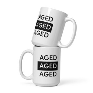 May include: Two white ceramic mugs stacked on top of each other. The mugs have black text that says "AGED" on them. The text is stacked vertically on the mugs.