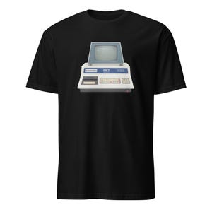 Może przedstawiać: Czarny t-shirt z grafiką retro komputera. Komputer ma niebieski ekran, a słowo "PET" jest widoczne. Koszulka ma klasyczny okrągły dekolt.