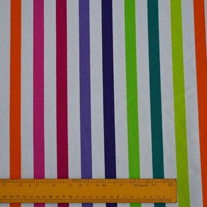 May include: A fabric with vertical stripes in various colors, including green, orange, pink, purple, and teal, against a white background. A yellow ruler is at the bottom of the image, measuring the fabric.