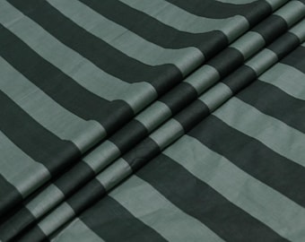 May include: Close-up of a fabric with a bold striped pattern. The stripes alternate between dark green and a lighter, muted green. The fabric is draped, showing folds and texture.