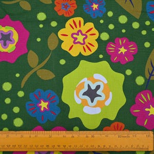 May include: A fabric with a repeating floral pattern. The design features large, stylized flowers in shades of pink, yellow, orange, and blue, set against a dark green background. The flowers have star-shaped centers and are accompanied by green leaves and dots. A ruler is at the bottom.