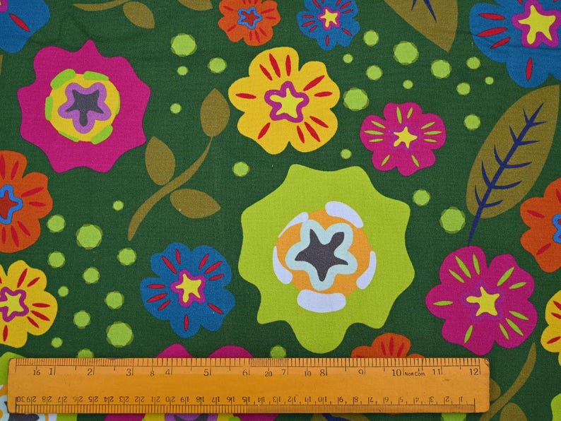 May include: A fabric with a repeating floral pattern. The design features large, stylized flowers in shades of pink, yellow, orange, and blue, set against a dark green background. The flowers have star-shaped centers and are accompanied by green leaves and dots. A ruler is at the bottom.