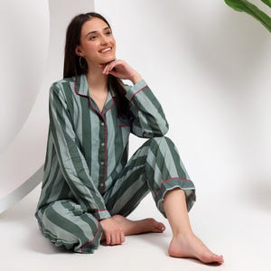 Op de afbeelding: Een pyjamaset met een pyjamashirt met lange mouwen en knopen en een bijpassende pyjamabroek met een verticaal gestreept patroon in afwisselende tinten donkergroen en saliegroen, afgewerkt met een dunne rode bies langs de kraag en manchetten.