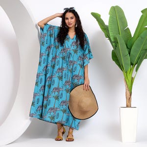 May include: A light blue kaftan with a white and black elephant print. The kaftan has a V-neck and a drawstring waist. The model is wearing the kaftan with a straw hat and sandals.