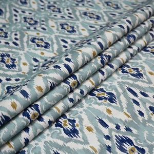 May include: Close-up of folded fabric with a repeating ikat pattern. The design features a mix of blue, white, and gold colors. The fabric has a soft, flowing appearance, with visible folds and creases.