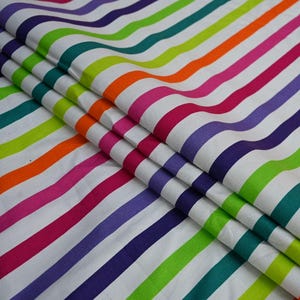 May include: Fabric with a repeating pattern of colorful, wavy stripes in shades of purple, pink, orange, green, and blue on a white background. The fabric is folded, showing the texture and layers of the material.