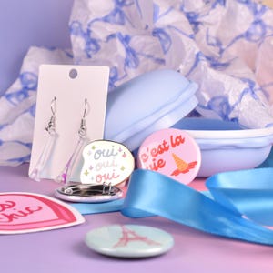 May include: A selection of pastel-coloured accessories, including earrings, pins, and a blue container. The earrings are clear with a purple accent. One pin says "Oui Oui Oui" and another says "C'est la vie". A blue ribbon is also present.