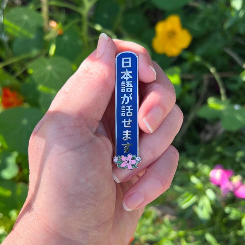 Peut inclure: &Eacute;pinglette &eacute;maill&eacute;e bleue et argent&eacute;e avec texte japonais. L'&eacute;pinglette pr&eacute;sente des caract&egrave;res japonais blancs sur fond bleu, avec un motif floral en bas. L'&eacute;pinglette est tenue dans une main sur un fond vert flou.