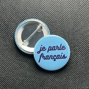 Może przedstawiać: Niebieski guzik z napisem "je parle français" w białym, ozdobnym stylu pisma.