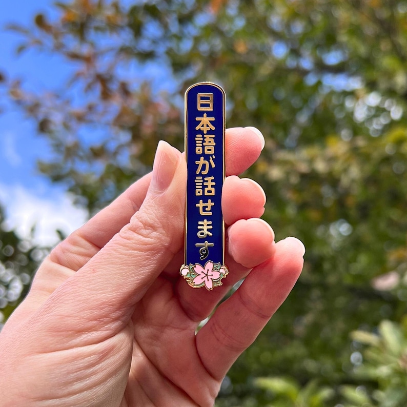 Peut inclure: Une &eacute;pingle en &eacute;mail bleu marine avec une bordure dor&eacute;e et du texte japonais. L'&eacute;pingle pr&eacute;sente un motif floral avec des accents roses et verts en bas. Le texte se lit "日本語が話せます".
