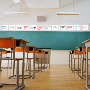Puede incluir: Un aula con una pizarra verde y filas de sillas naranjas y plateadas. La pizarra tiene varios carteles coloridos con frases en espa&ntilde;ol, incluyendo "Yo no s&eacute;", "Presente", "No lo tengo", "&iquest;C&oacute;mo?" y "Necesitamos m&aacute;s tiempo".