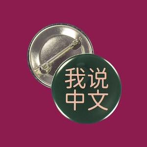 Może przedstawiać: Zielony guzik z szpilką z tyłu. Guzik ma różowy napis "Mówię po chińsku" w chińskich znakach.