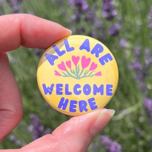 Puede incluir: Un botón amarillo con las palabras "ALL ARE WELCOME HERE" en letras azules. El botón también presenta un diseño floral con corazones rosas y tallos verdes. El botón está sostenido en una mano.