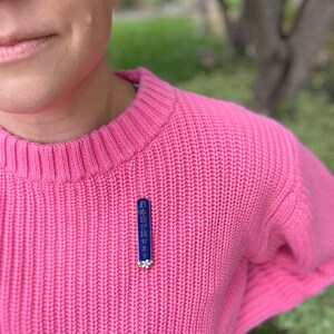 Peut inclure: Un pull rose tricot&eacute; avec une &eacute;pingle bleue et blanche. L'&eacute;pingle porte du texte japonais et un petit motif floral. Le pull est de couleur rose vif unie.