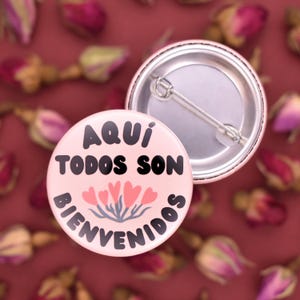 May include: Pink button with the text "Aqui Todos Son Bienvenidos" in black lettering. The button has a floral design with red hearts.