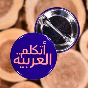 May include: Two round, blue pinback buttons. One button displays the text "أَتَكَلَّمُ الْعَرَبِيَّةُ" in pink. The other button shows the pinback. The background is a light brown, wooden surface.