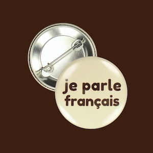 Könnte beinhalten: Ein weißer Knopf mit einer braunen Aufschrift, die "je parle français" lautet. Der Knopf hat eine silberne Rückseite mit einer Sicherheitsnadel.