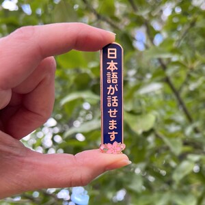 Peut inclure: &Eacute;pingle rectangulaire en &eacute;mail avec un fond bleu marine et une bordure dor&eacute;e. L'&eacute;pingle pr&eacute;sente du texte japonais en rose et un motif de fleur de cerisier rose en bas. L'&eacute;pingle est tenue entre deux doigts sur un fond vert flou.