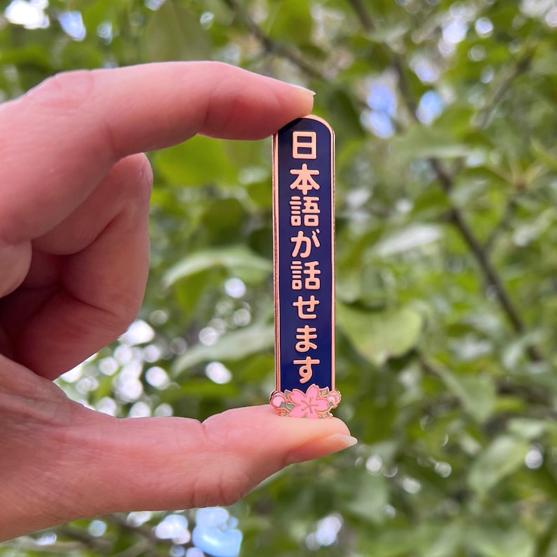 Peut inclure: &Eacute;pingle rectangulaire en &eacute;mail avec un fond bleu marine et une bordure dor&eacute;e. L'&eacute;pingle pr&eacute;sente du texte japonais en rose et un motif de fleur de cerisier rose en bas. L'&eacute;pingle est tenue entre deux doigts sur un fond vert flou.