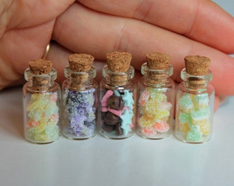 May include: Five small glass jars with cork lids filled with colorful candies. The jars are arranged in a row on a white surface.