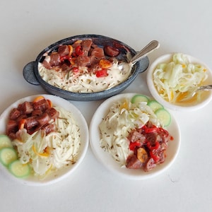 May include: Miniature food set featuring a dark gray oval dish with a creamy base and meat, two white plates with pasta, meat, and cucumber slices, and a small bowl of salad. A small spoon rests in the oval dish.