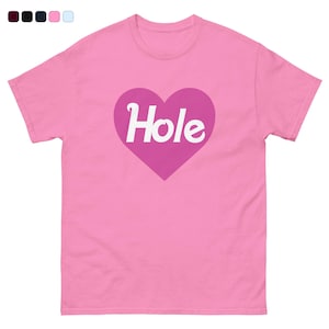 May include: A pink t-shirt featuring a large, heart-shaped graphic in a darker shade of pink. The word "Hole" is written in white, cursive font inside the heart. The shirt has a crew neck and short sleeves.
