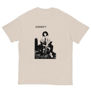 May include: A beige t-shirt featuring a black and white image of a person sitting on a chair with a microphone and the word "COSEY" printed on the front.