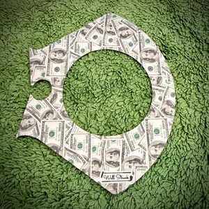 Puede incluir: Un recorte decorativo con tem&aacute;tica de dinero con un gran agujero central, estampado con im&aacute;genes de billetes de 100 $. El recorte es blanco con un borde negro y el texto "Will Struck" en la parte inferior. El fondo es una superficie verde texturizada.