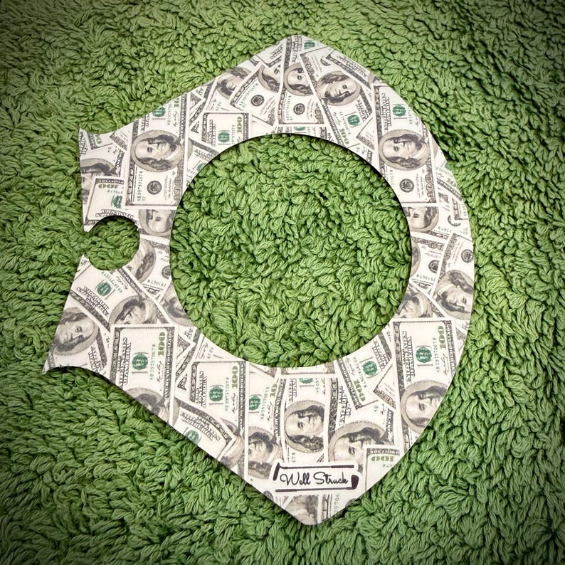 Puede incluir: Un recorte decorativo con tem&aacute;tica de dinero con un gran agujero central, estampado con im&aacute;genes de billetes de 100 $. El recorte es blanco con un borde negro y el texto "Will Struck" en la parte inferior. El fondo es una superficie verde texturizada.