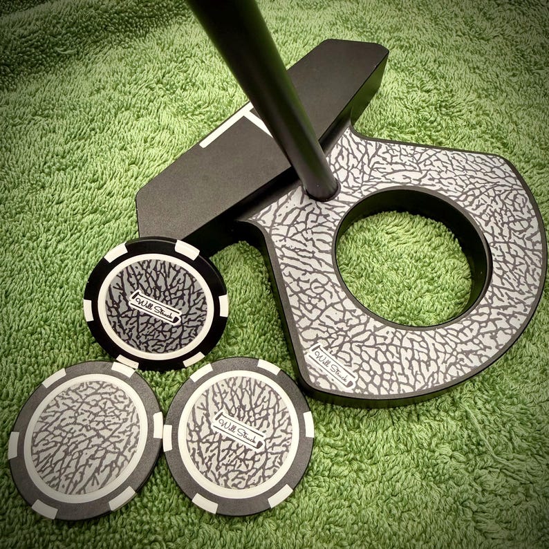 Pu&ograve; includere: Un putter da golf nero e grigio con un motivo unico a pelle di elefante, insieme a tre segnapunti da golf abbinati. I segnapunti e il putter hanno il testo "Will Stacks". Il putter &egrave; su una superficie verde.