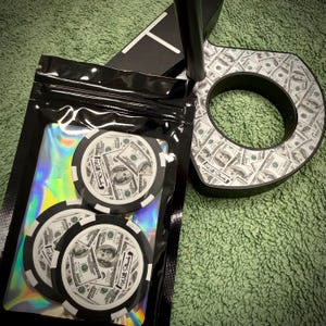 Puede incluir: Fichas de p&oacute;quer con un dise&ntilde;o de billetes de 100 $ y el texto "Will Stack" se muestran en una bolsa de pl&aacute;stico transparente. Tambi&eacute;n se ve una funda negra para la cabeza del palo de golf con un dise&ntilde;o de billete de d&oacute;lar. Los art&iacute;culos est&aacute;n sobre una superficie verde.