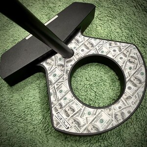 Puede incluir: Putter de golf negro con un dise&ntilde;o tem&aacute;tico de dinero. La cabeza del putter presenta un patr&oacute;n de billetes de cien d&oacute;lares. El putter tiene un eje negro y una abertura circular en la cabeza. Las palabras "Will Struck" est&aacute;n impresas en el putter.